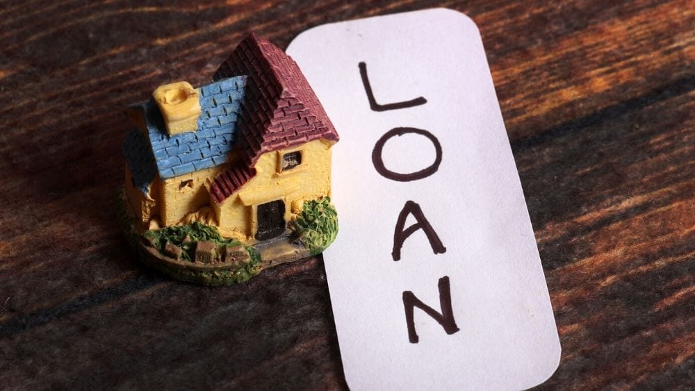 A small figurine of a house with a red and blue roof and yellow walls next to a piece of paper that says "loan" in capital letters.