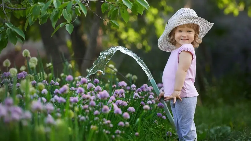 a young girl in a white hat holds a hose and is watering plants in a garden