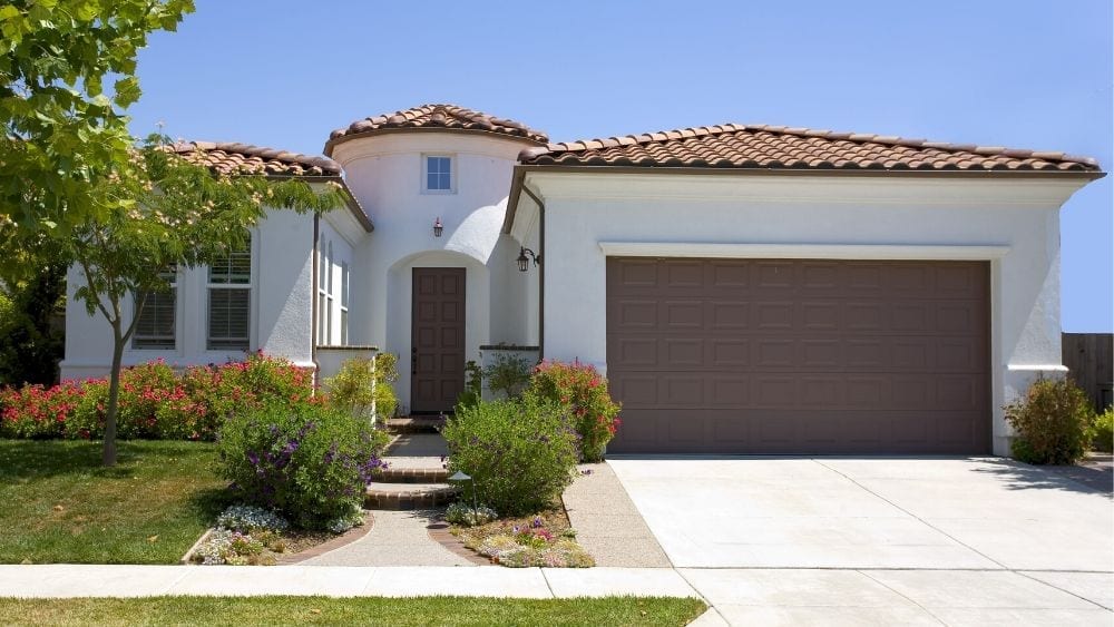 A two-story Spanish-style home with white stucco walls, red barrel tile roof, and a brown garage door and brown front door.