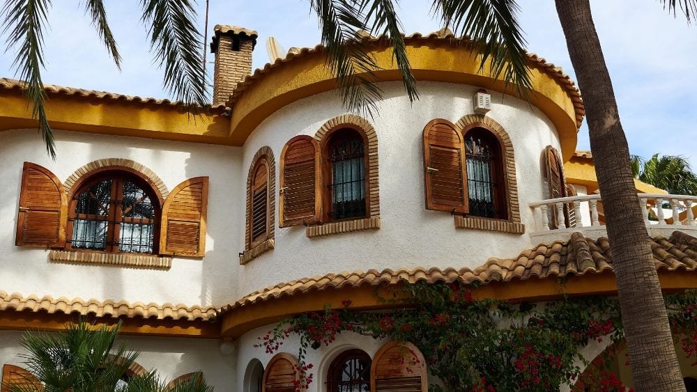 The upper level exterior of a home with stucco walls, warm yellow trim, and arched windows with stone lining and muted yellow shutters.