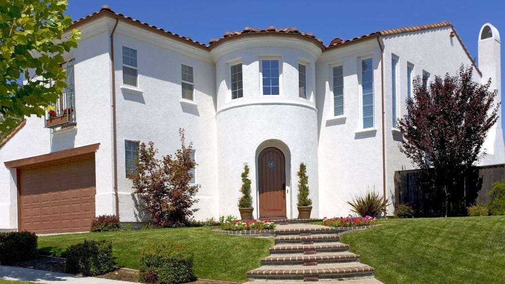 A large Spanish-style home with white stucco exterior walls and many windows. The front door is round, and it and the garage door are orange-brown. Stone steps lead through a manicured lawn to the front door.