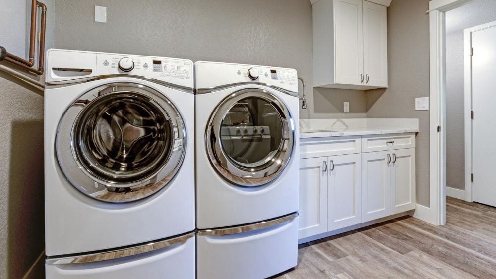 A taupe laundry room with a white washer and dryer, sink, and white cabinets.