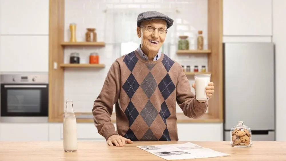An older man standing at the kitchen counter, smiling and holding a glass of milk.