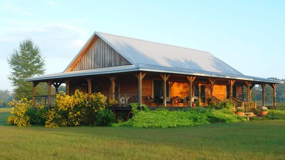 A log cabin with a metal roof and a wraparound porch surrounding by shrubs and pasture