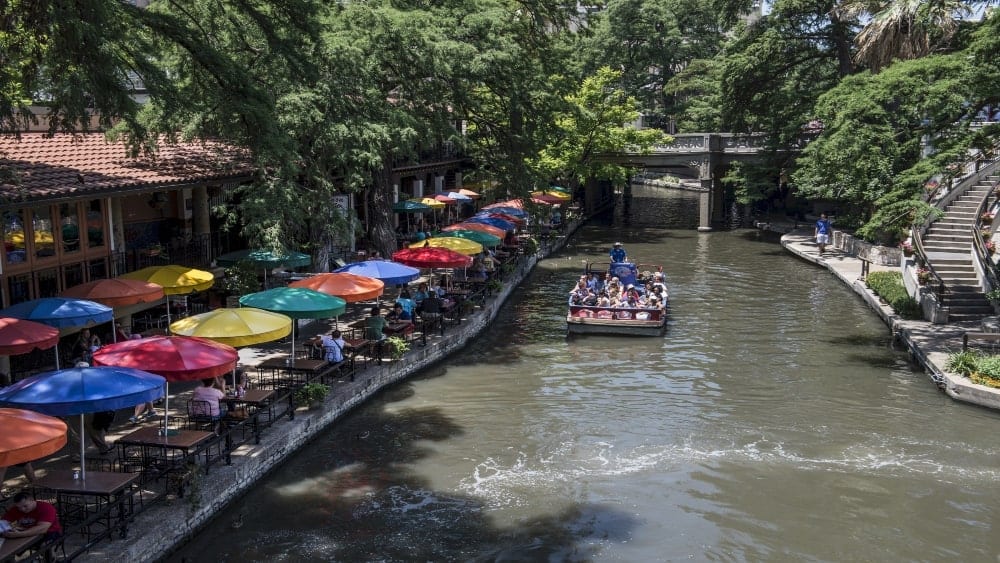 View of the San Antonio River in San Antonio Texas with a riverboat filled with tourists on the water and brightly colored umbrellas lining the River Walk on the left