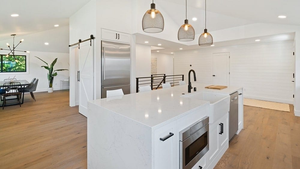White modern farmhouse kitchen with silver appliances, pendant lights, and brown hardwood flooring.