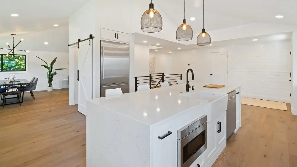 White modern farmhouse kitchen with silver appliances, pendant lights, and brown hardwood flooring.