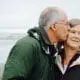 Senior man kissing wife on the cheek with a beach in the background.