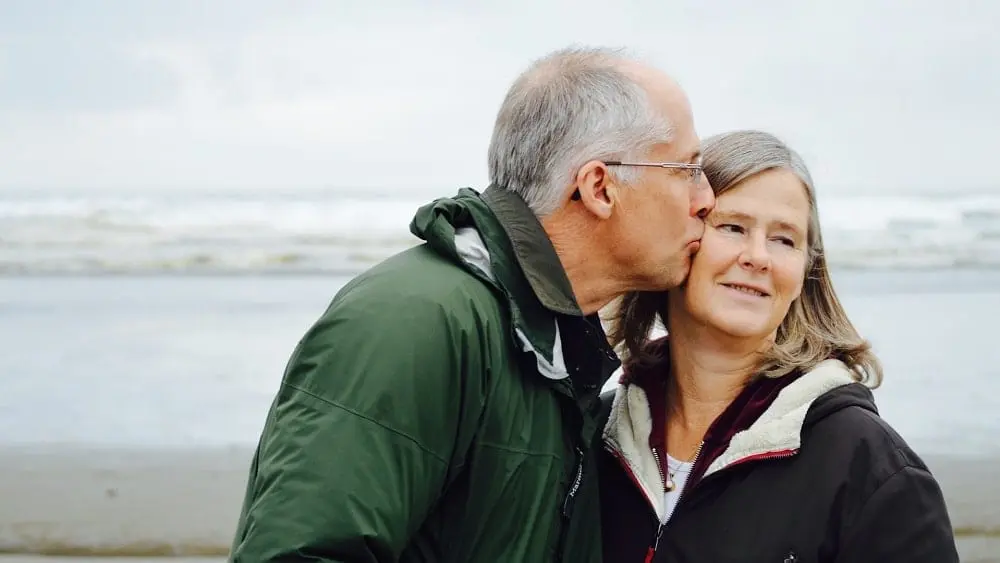 Senior man kissing wife on the cheek with a beach in the background.