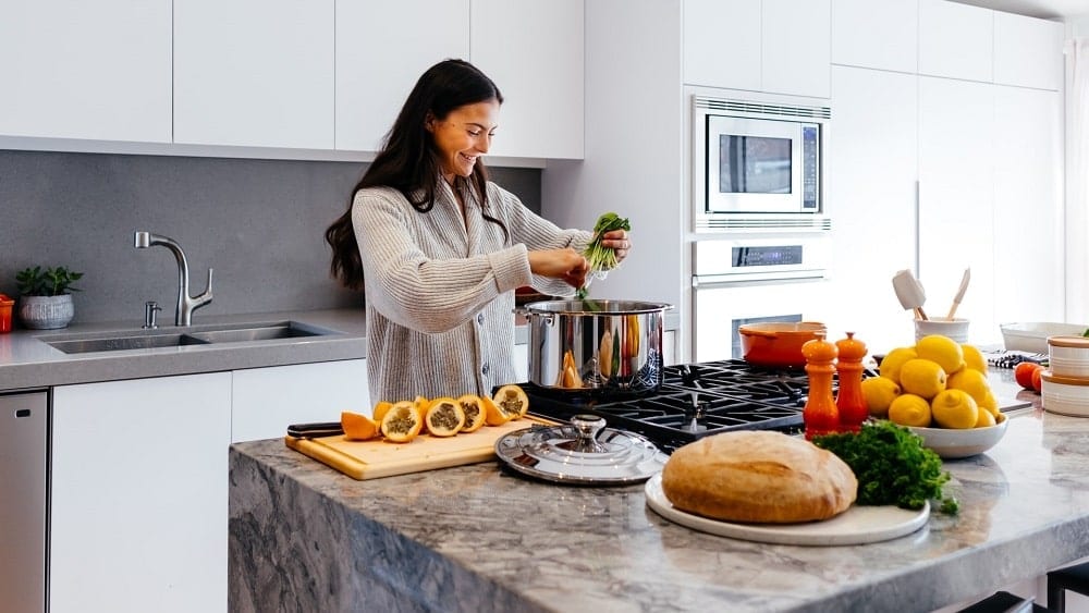 Woman in modern kitchen standing next behind marble island and preparing food.