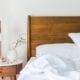 Bed with wooden headboard and white sheets with a wooden nightstand holding copper lamp, potted plants, and small accessories.