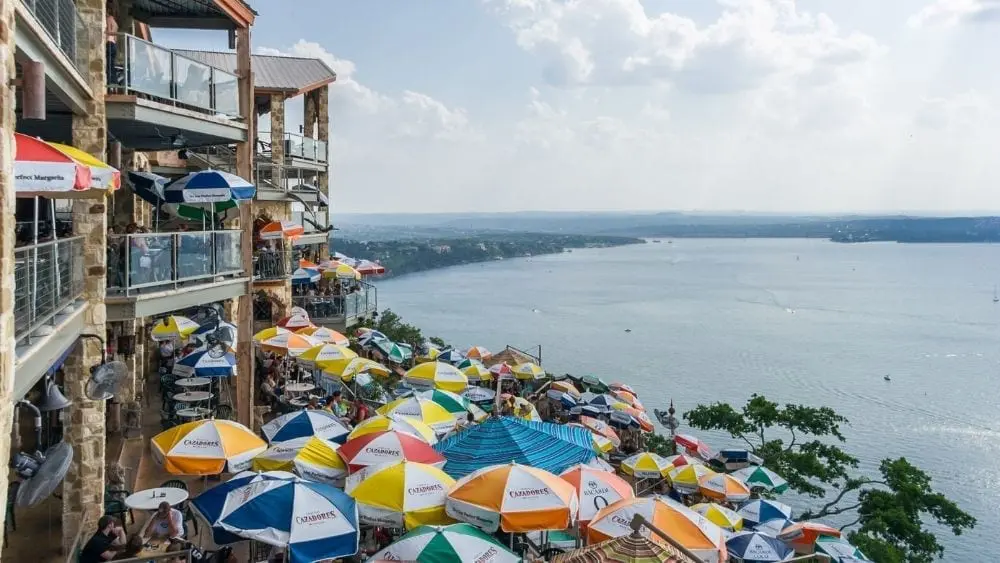 Patio tables and umbrellas in front of a multi-story building, looking over Lake Travis.