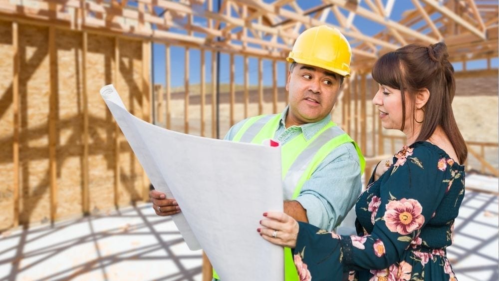 A man in a construction hat holding designs of a home for a woman to review. They're standing in the frame of a new home.