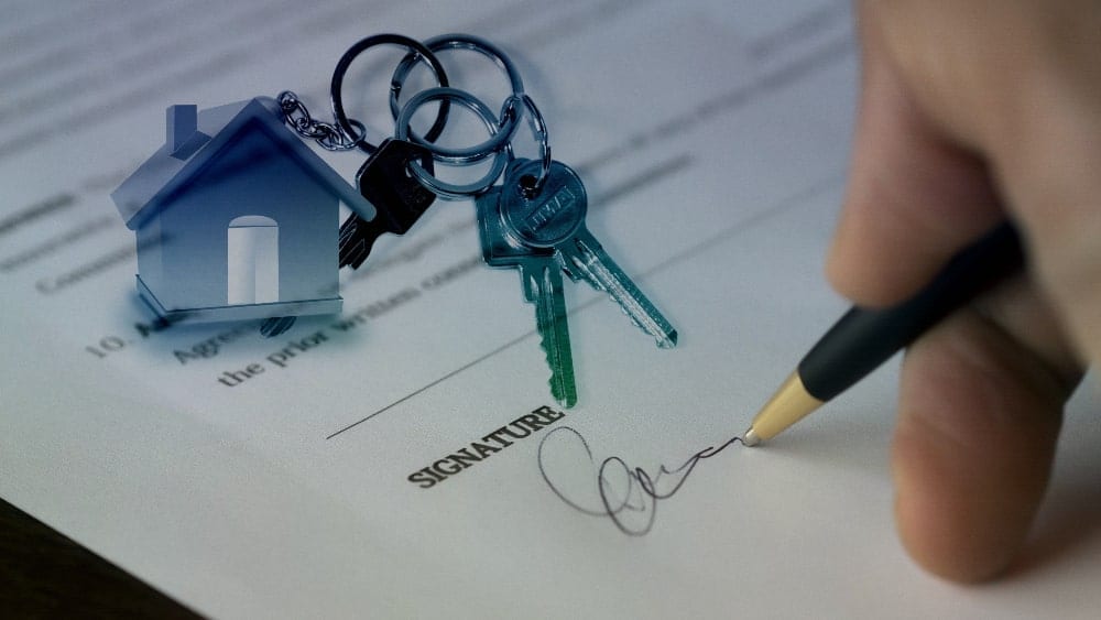 A house keychain with two keys sits on top of a contract with a hand signing the document
