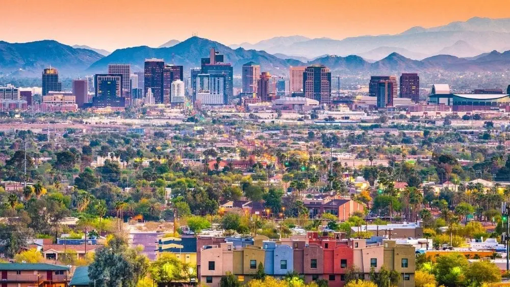 The Phoenix skyline against a mountain range and orange sky, with colorful buildings and houses in the foreground.
