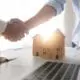 Close-up of a handshake over the design plans of a home, a computer, and construction hats.