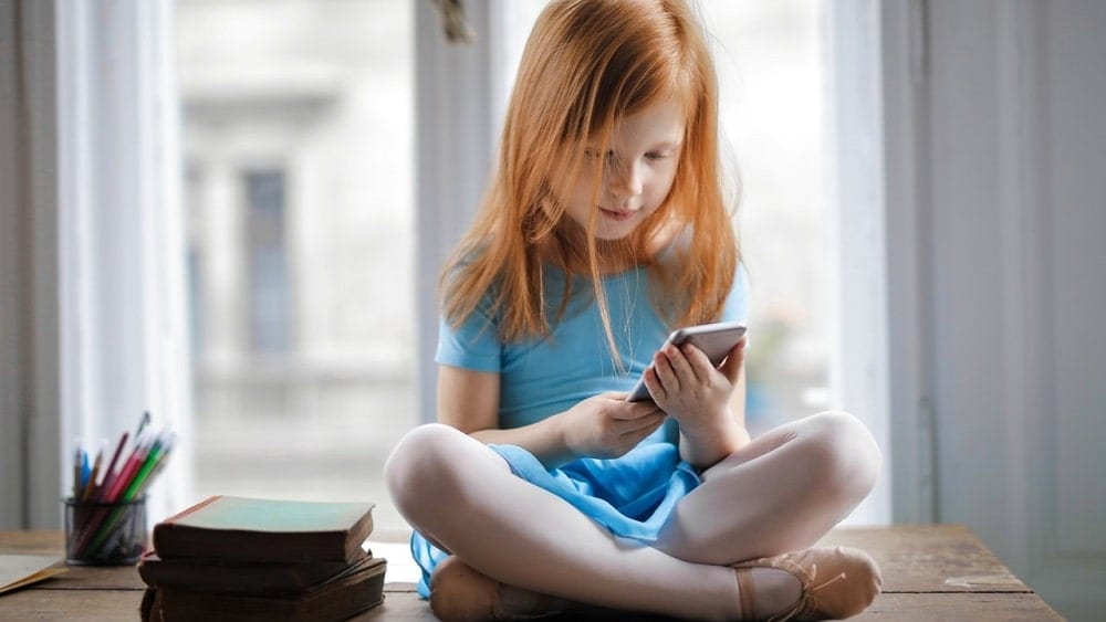 Young girl sitting on desk next to books holding a smartphone.