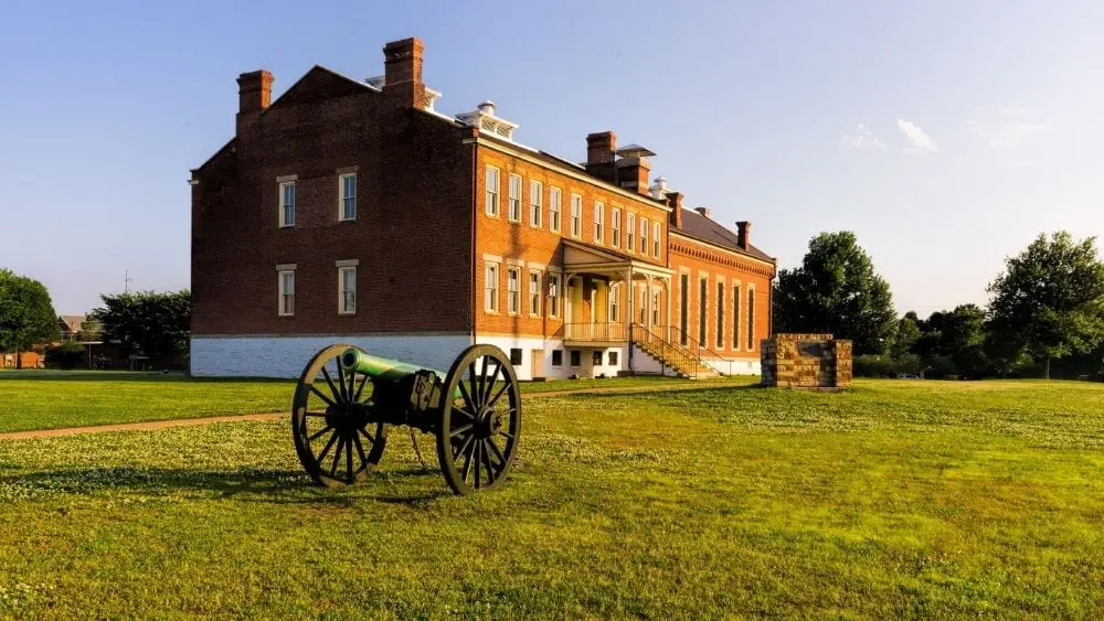 Angled view of an old cannon on wheels in front of a large brick building.