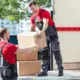 two movers in red and black uniforms move boxes from the back of a moving truck