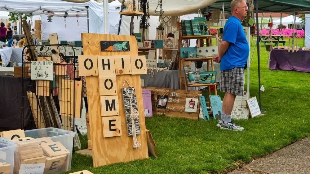 An outdoor market of artisan goods; close to the front is a sign of large Scrabble tiles reading "Ohio" and "Home" with the "h" overlapping.