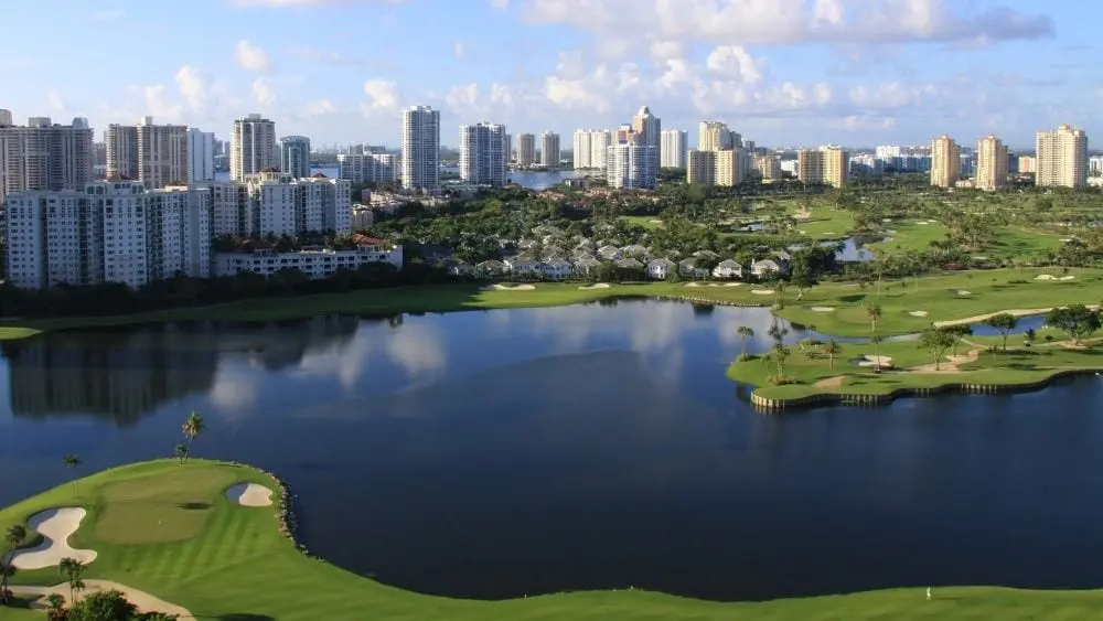 Aerial view of a lake surrounded by a well-maintained golf course. A city skyline stands in the background.