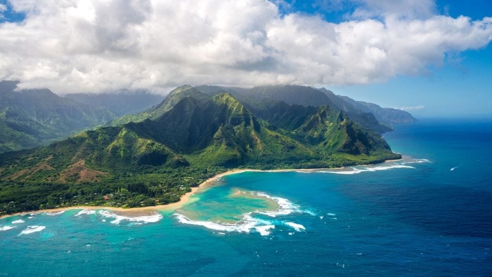 Drone image of Kauai Island surrounded by blue waters and covered with greenery.