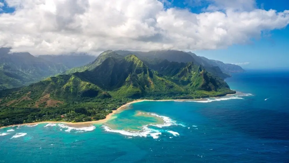 Drone image of Kauai Island surrounded by blue waters and covered with greenery.