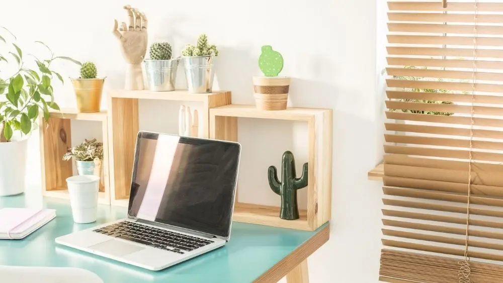 A blue desk with a laptop and bamboo trinkets and blinds on the window.