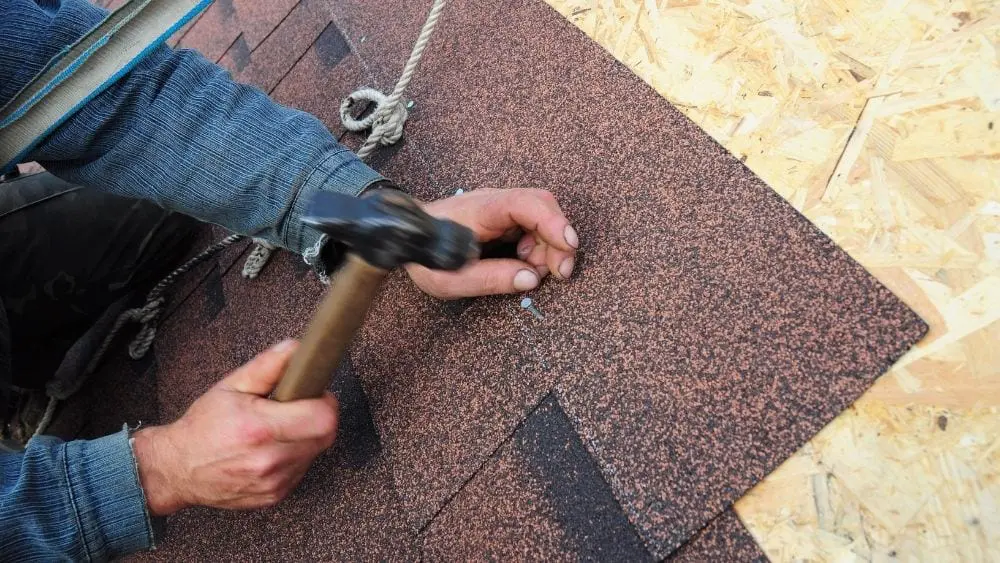 Close-up of someone hammering in asphalt shingles to a roof.