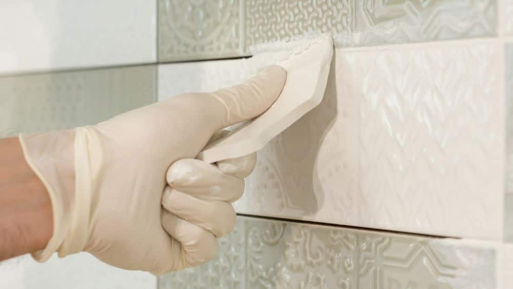 Close-up of someone installing white and grey tile and sealing the edges with grout.