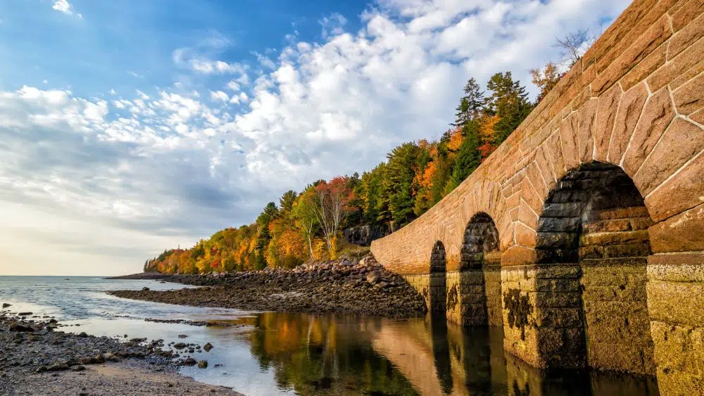 View from a rocky beach looking up at a stone bridge with arches. At the end of the bridge is a forest with fall-colored trees.