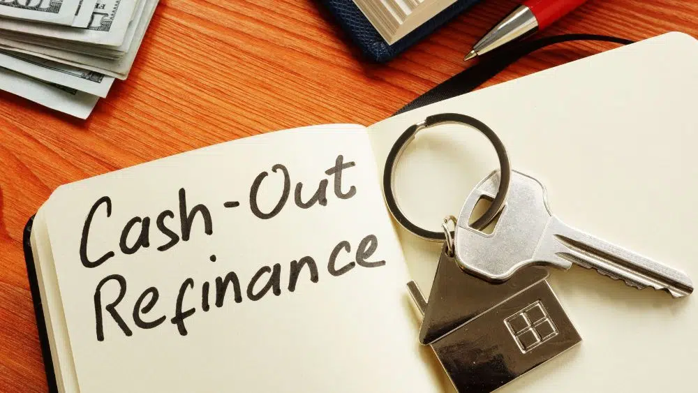 An open notebook with "cash-out refinance" written on the page. A stack of money, pen, and silver house keychain with a key are in the image.