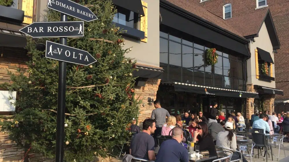 People sitting on a patio outside of a restaurant. A christmas tree is on one side, and a pole with various metal signs pointing to different bars is in front of it.