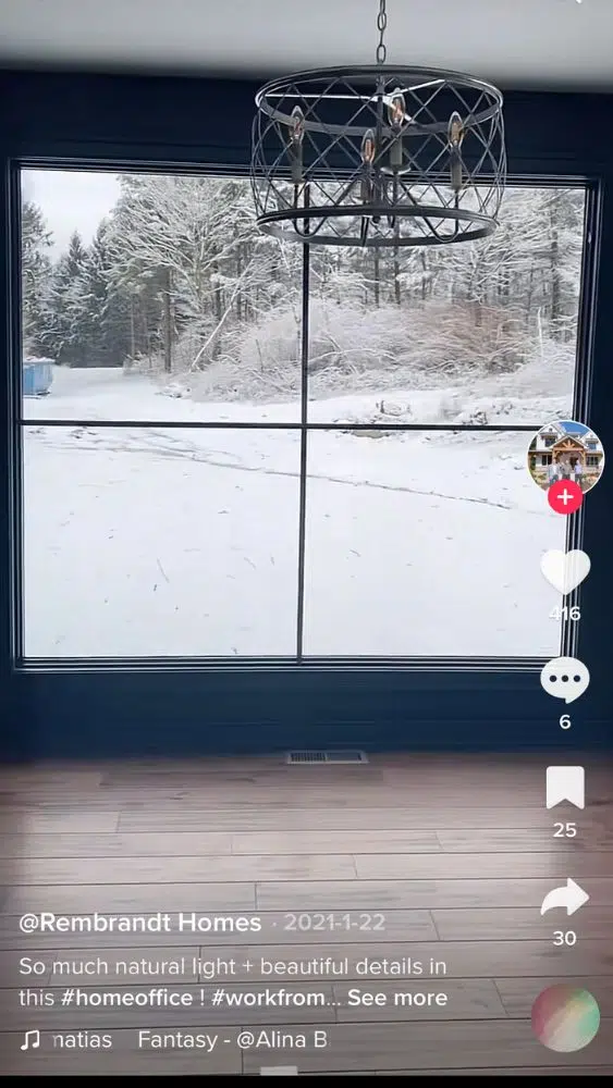Screenshot of @rembrandthomes' TikTok video showing a floor-to-ceiling window with a modern chandelier hanging in front of it. There is a snowy landscape outside.