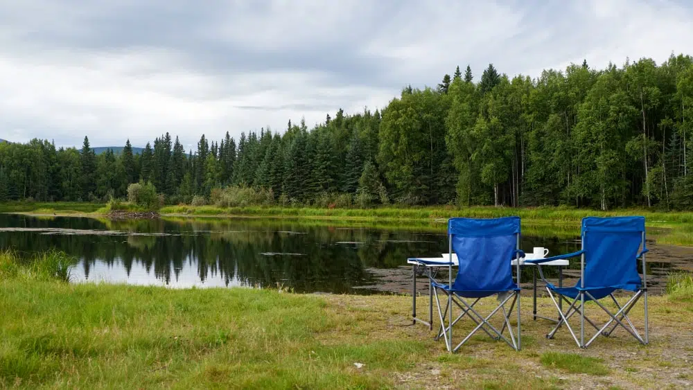 Two blue camping chairs with table and coffee cups in front of small lake with woods in the background, Chena River State Recreation Area, Alaska