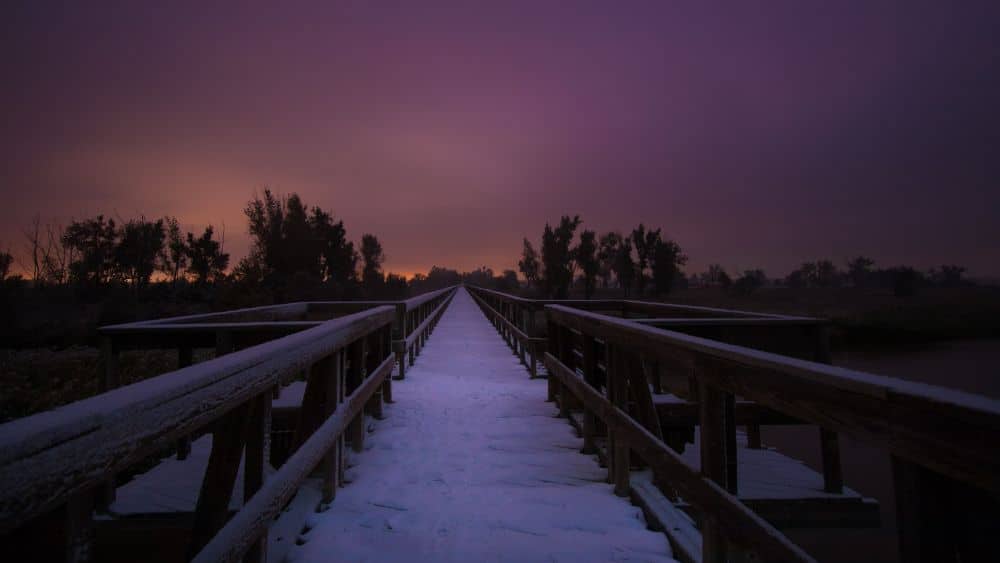 View looking down a snow-covered path that disappears into the horizon. The sun is setting, making the sky orange and purple.