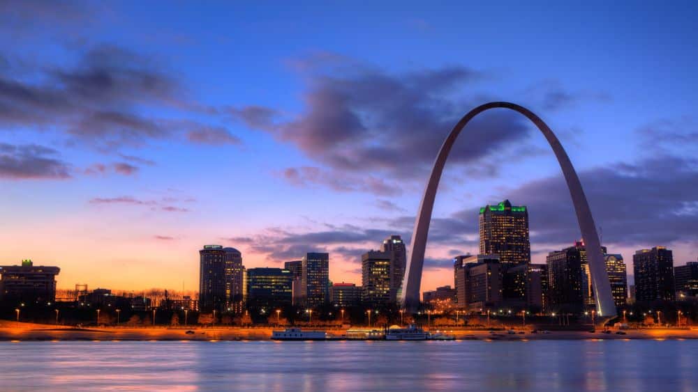 City skyline at sunset with lit-up buildings and a large metal arch towering above them all.