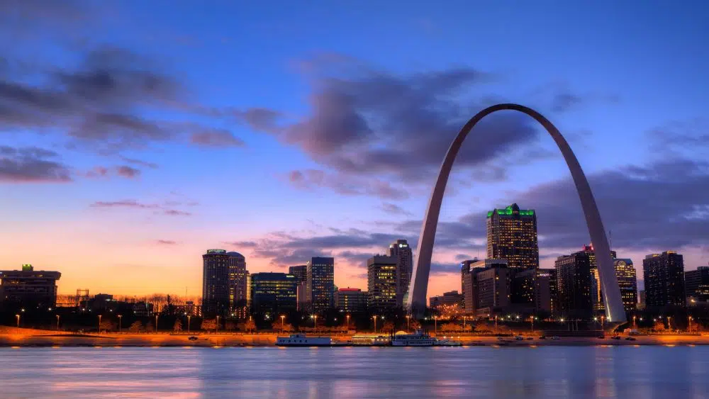 City skyline at sunset with lit-up buildings and a large metal arch towering above them all.