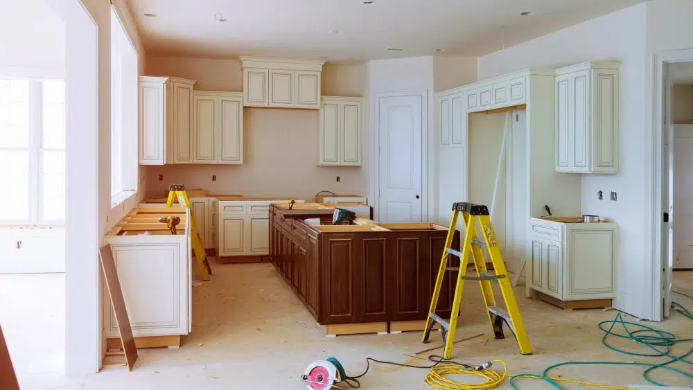 A work-in-progress kitchen renovation showing new white cabinets along the walls and dark brown cabinets for an island, with tools lying around and a yellow stepladder in the foreground.