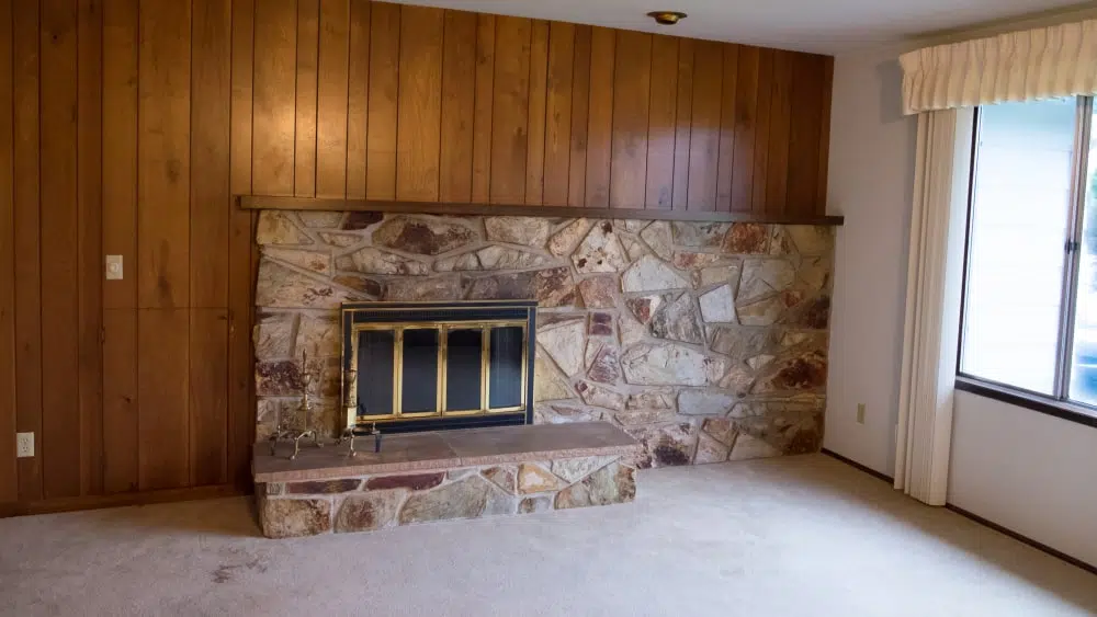 A 1970s-style living room with old beige carpet, stone fireplace with brass surround, and wood paneling on the wall and old cream curtains on the window