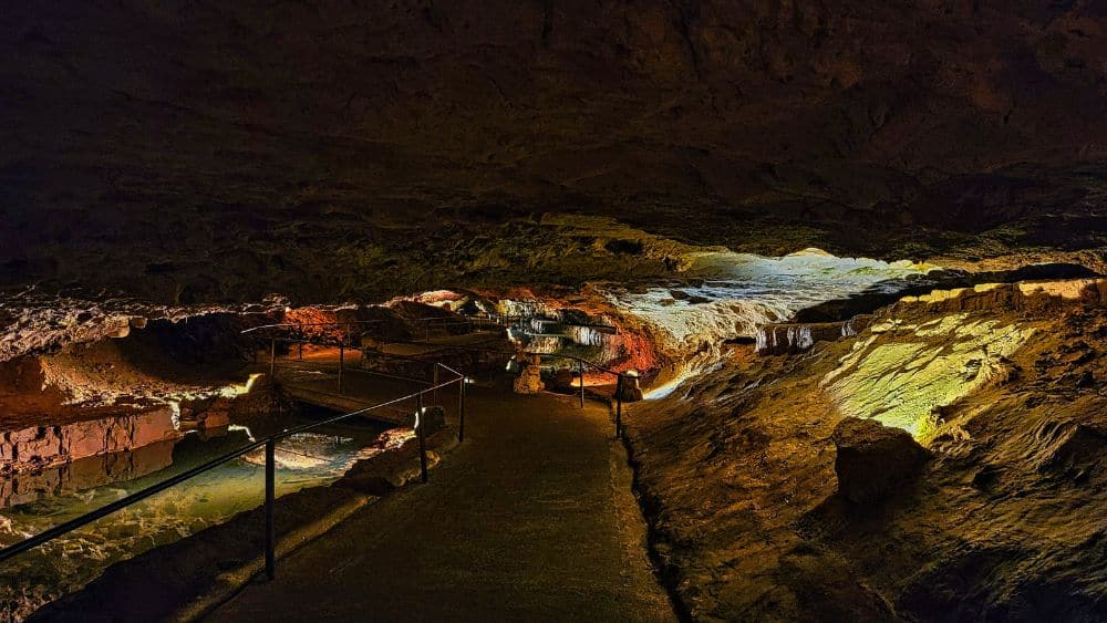 A dark cavern with a walkway down the middle. Sections of the cavern are lit in yellow lights, showing off jagged formations. Toward the bottom left is a small pool of water.