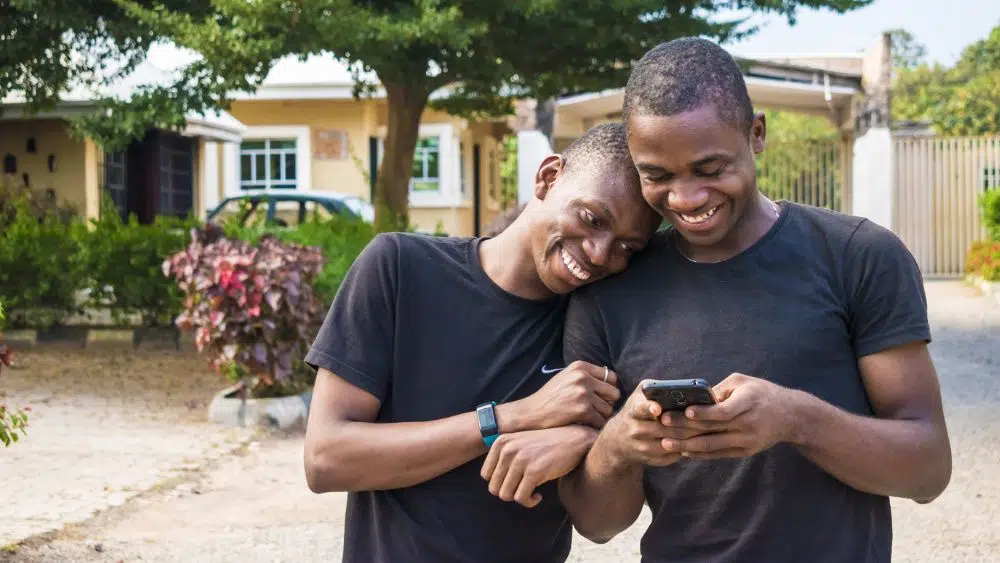 Two masculine people of color standing in front of a home, smiling down as they look at a phone. One person has their head resting on the shoulder of the other.