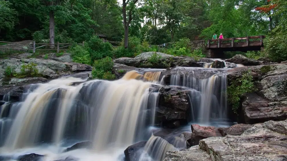 scenic view of the waterfall at Devil's Hopyard State Park, with two people standing on a viewing bridge in the upper righthand corner of the image
