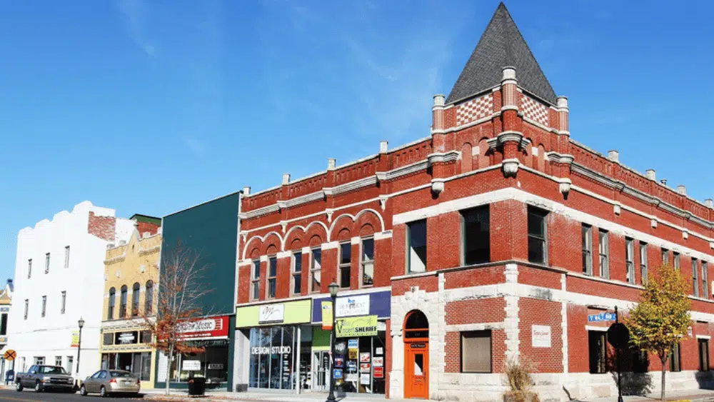 A red brick building on the corner of Walnut and Main Street in downtown Kokomo. To the left of the building is local shops with parked cars and a street lamp with signs pointing to street names.
