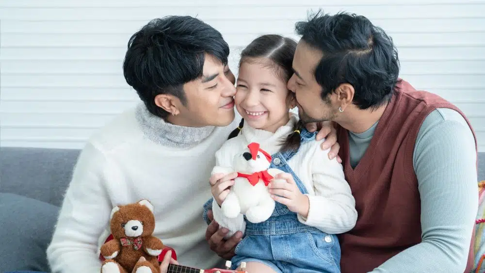 A smiling family made up of two masculine people of color and a child, all smiling as they sit on a couch together. The child is holding a stuffed bear.