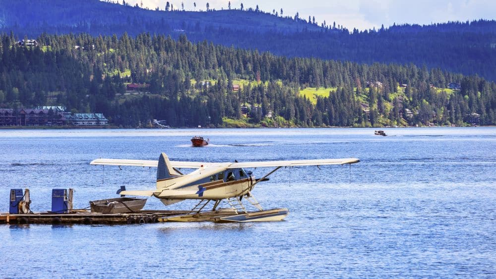 A small water plane floating next to a pier in a body of water. Mountains covered in trees are in the background.