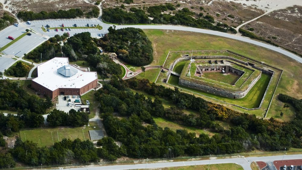 Aerial view of a historic fort. There is a brick building with a white roof on the lefthand side, and the fort itself is a pentagon dropped into the ground.