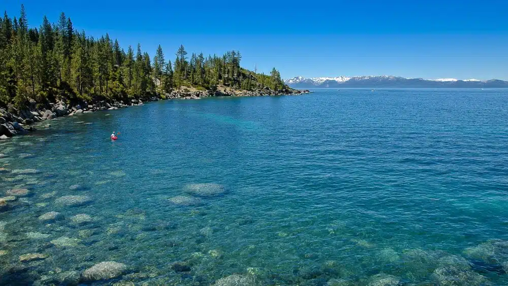 Clear blue lake with a rocky shore covered in trees and snowy mountains in the distance.