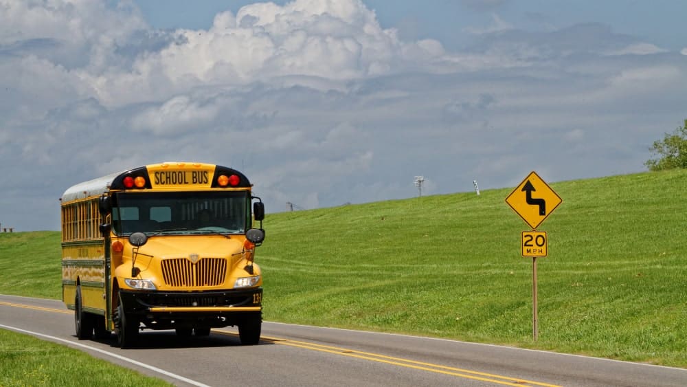 Image of a yellow school bus on a two-lane road and to the right is a yellow road size that says 20 mph