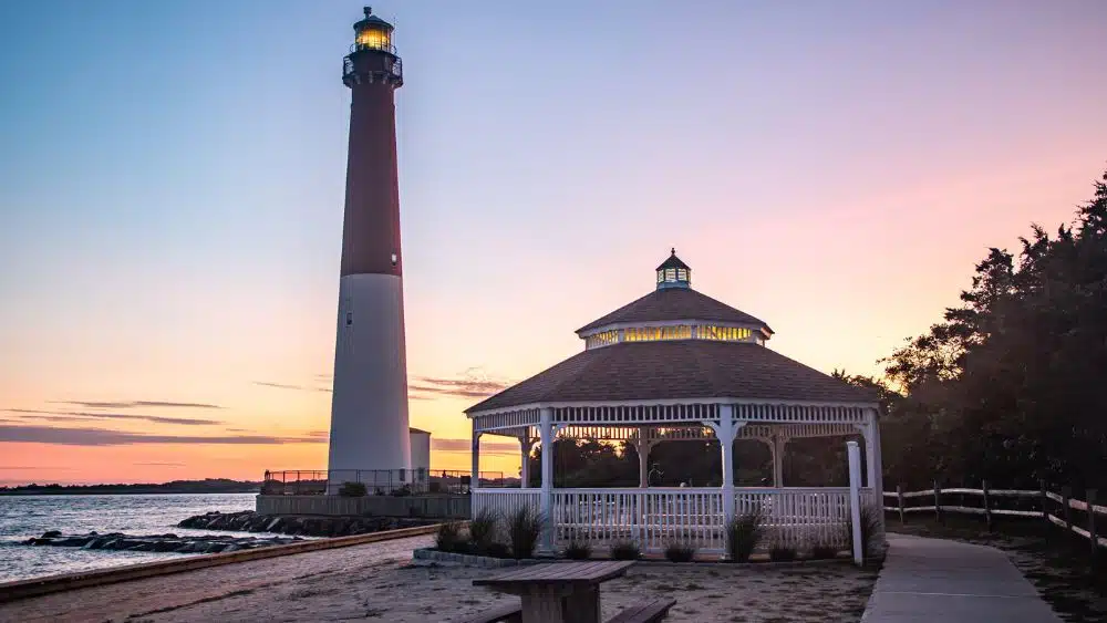 Red and white lighthouse next to a gazebo at sunset.